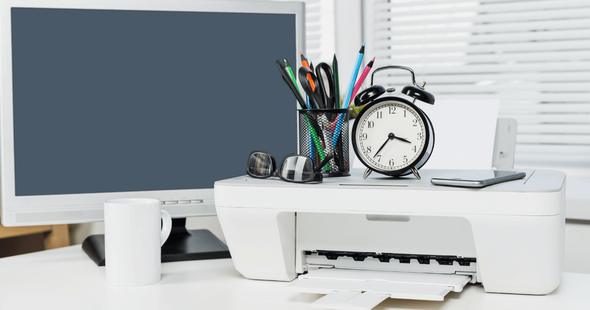 Desk setup with printer, clock, and computer in a bright office environment during daytime work hours
